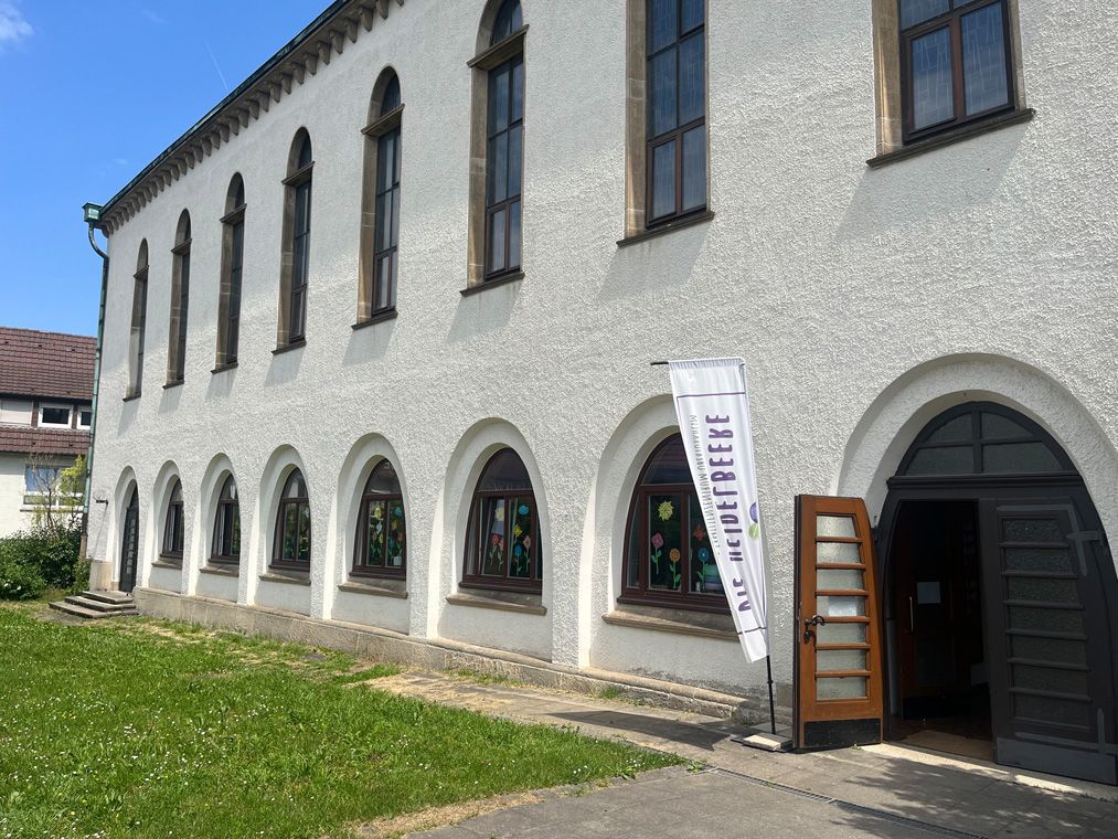 Außenansicht eines weißen Gebäudes mit Rundbogenfenstern und Fahnenbanner am Eingang.