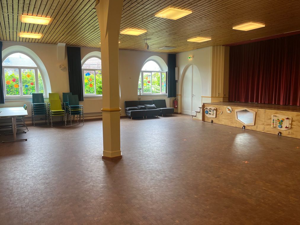 Saal mit Bühne, Stühlen und dekorierten Fenstern mit bunten Blumenbildern.