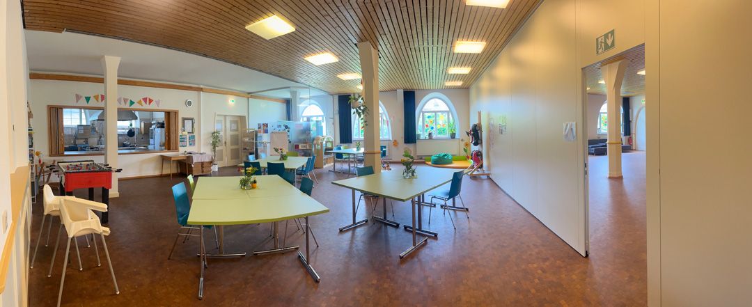Panoramaaufnahme eines Raumes mit grünen Tischen, Stühlen und Küchenbereich.
