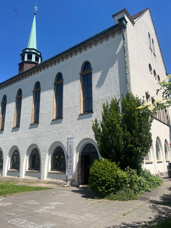 Gebäude mit Turm und weißen Rundbogenfenstern, davor ein Hof mit Hüpfspiel in Kreide.